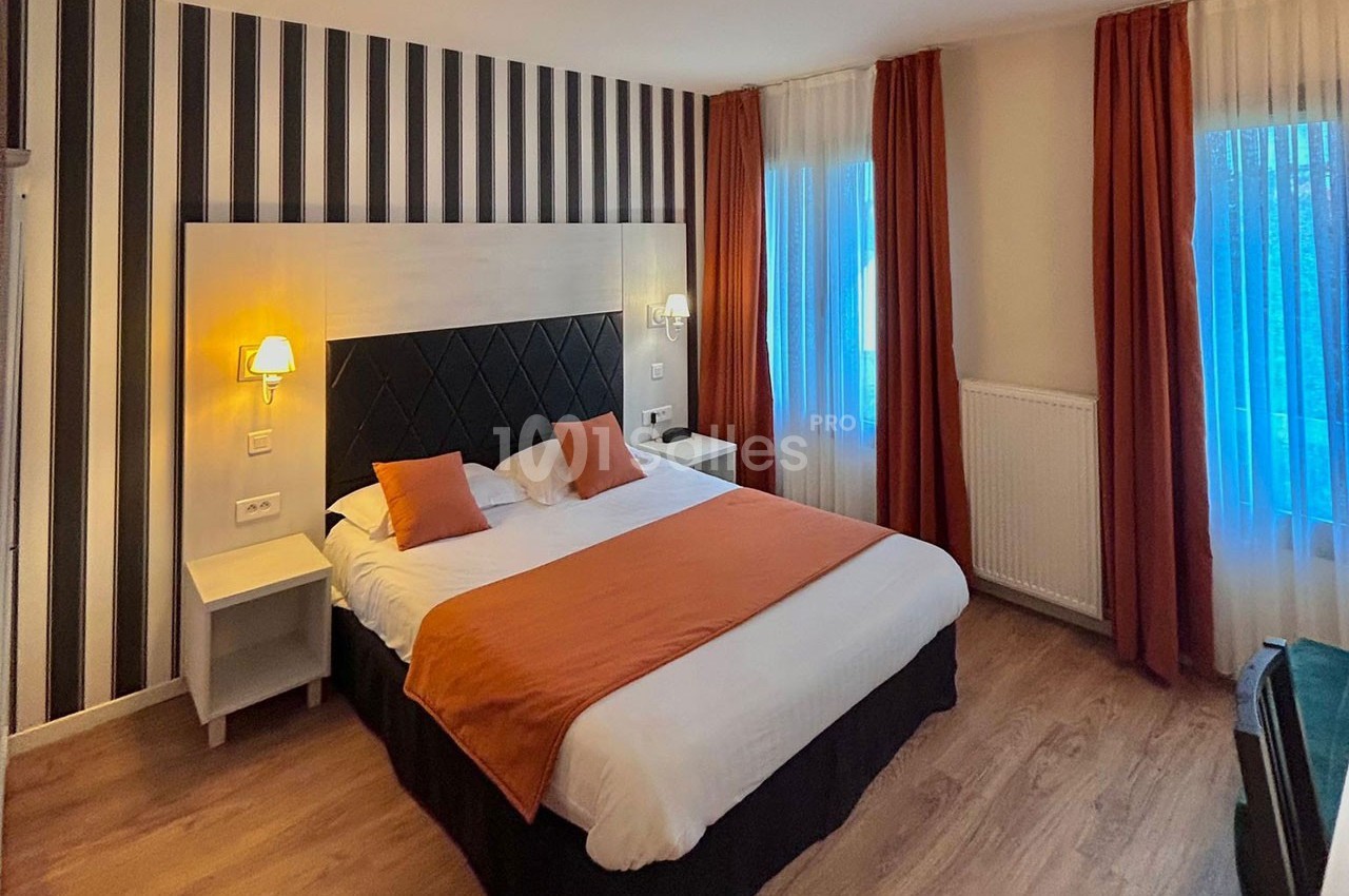 Chambre d'hôtel avec lit double, tête de lit rembourrée, murs à rayures, rideaux orange et éclairage doux.