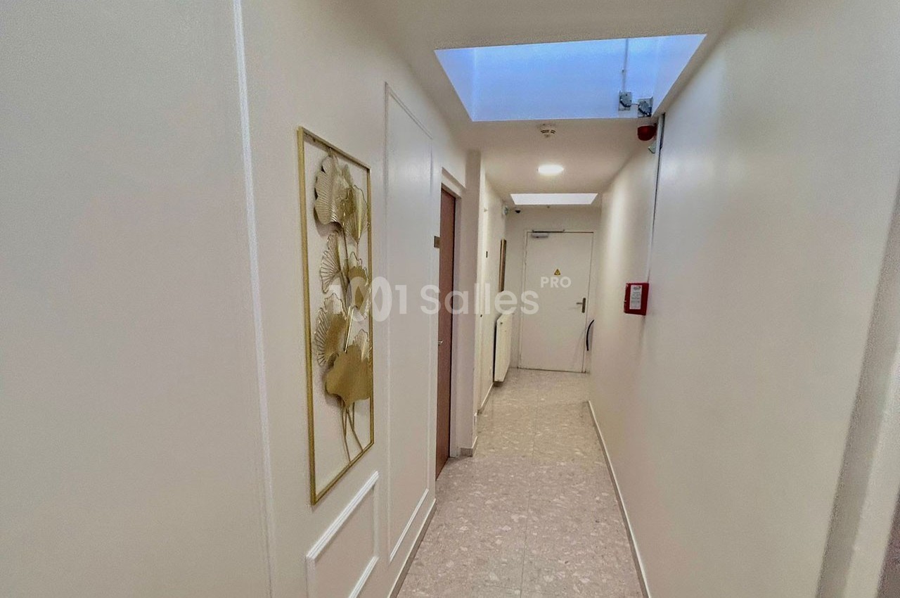 Couloir lumineux avec murs blancs, tableau floral encadré à gauche, porte au fond et éclairage naturel par une lucarne.