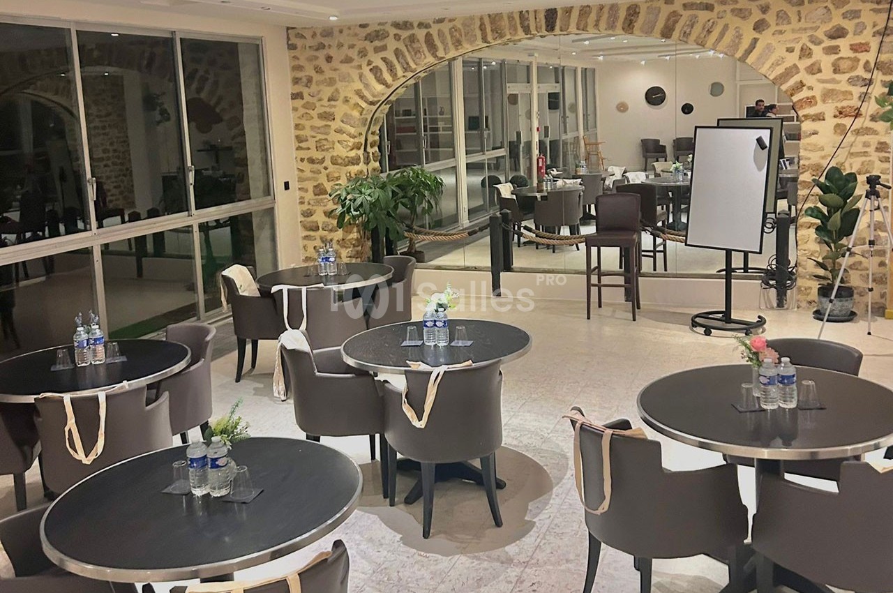 Salle lumineuse avec tables rondes, chaises en cuir, murs en pierre apparente et tableau blanc en arrière-plan.