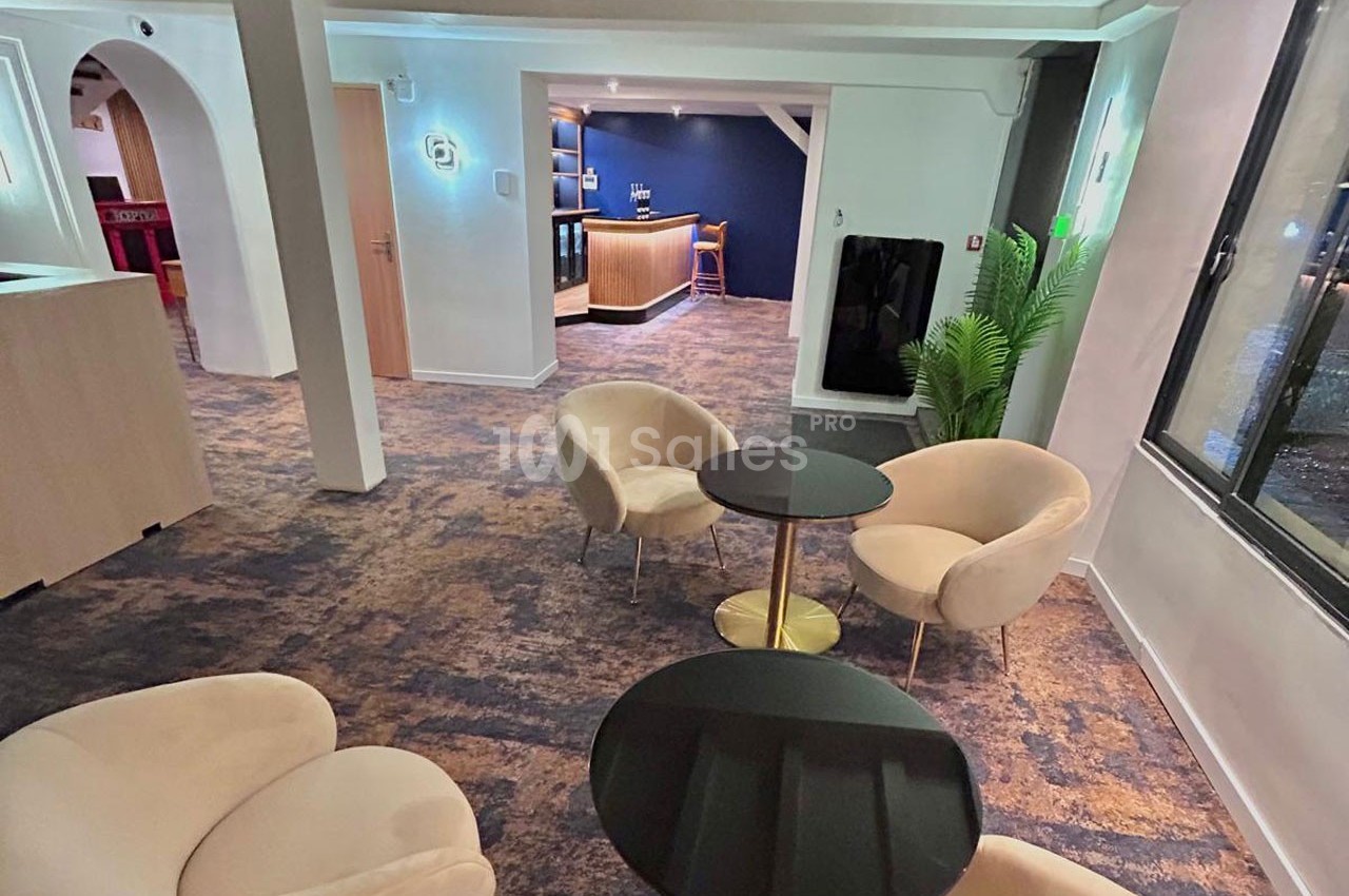 Espace intérieur avec fauteuils beiges, tables rondes noires, tapis bleu et comptoir de bar en arrière-plan.