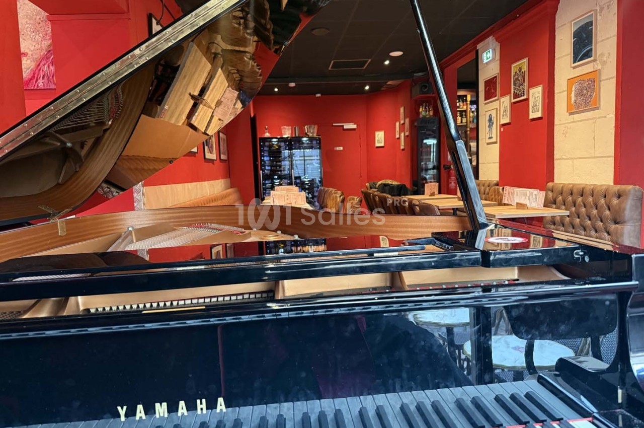 Piano à queue noir dans un restaurant aux murs rouges, avec des tables dressées et des fauteuils en cuir.