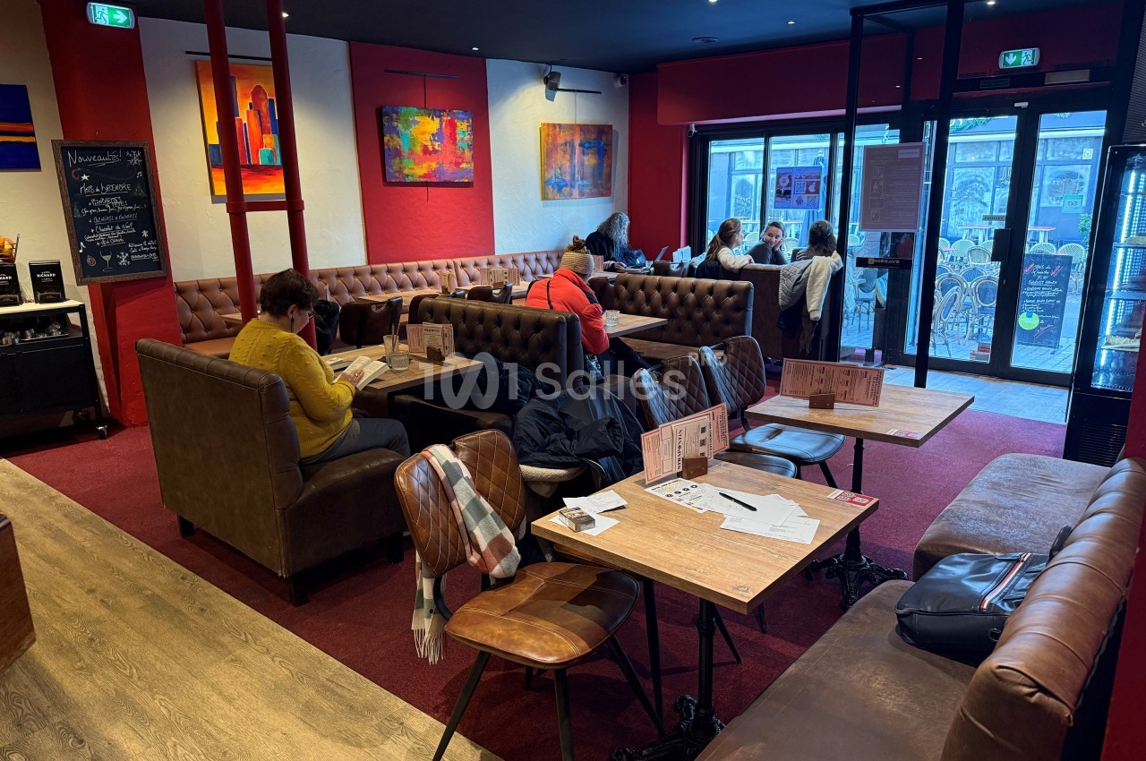 Intérieur d'un café avec des tables en bois, des banquettes et des clients assis, certains lisant ou discutant.