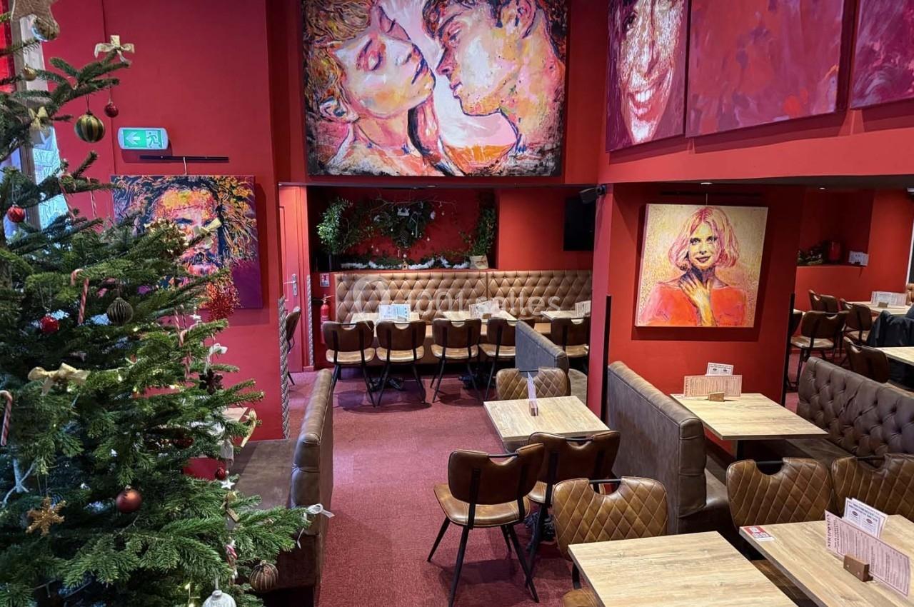 Salle de restaurant aux murs rouges décorée de peintures colorées, avec des tables en bois et un sapin de Noël à gauche.