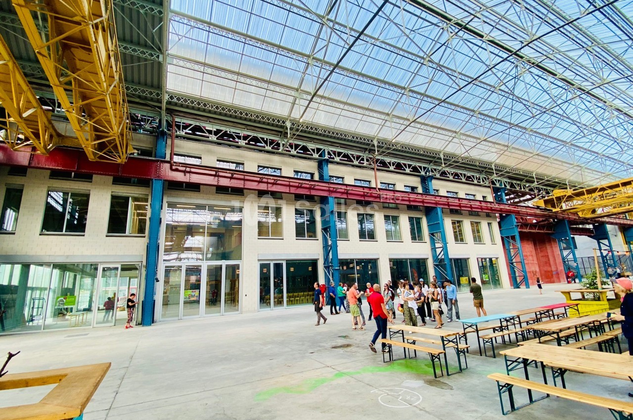Vue intérieure d'un grand bâtiment industriel rénové avec des structures métalliques, des tables et des visiteurs.