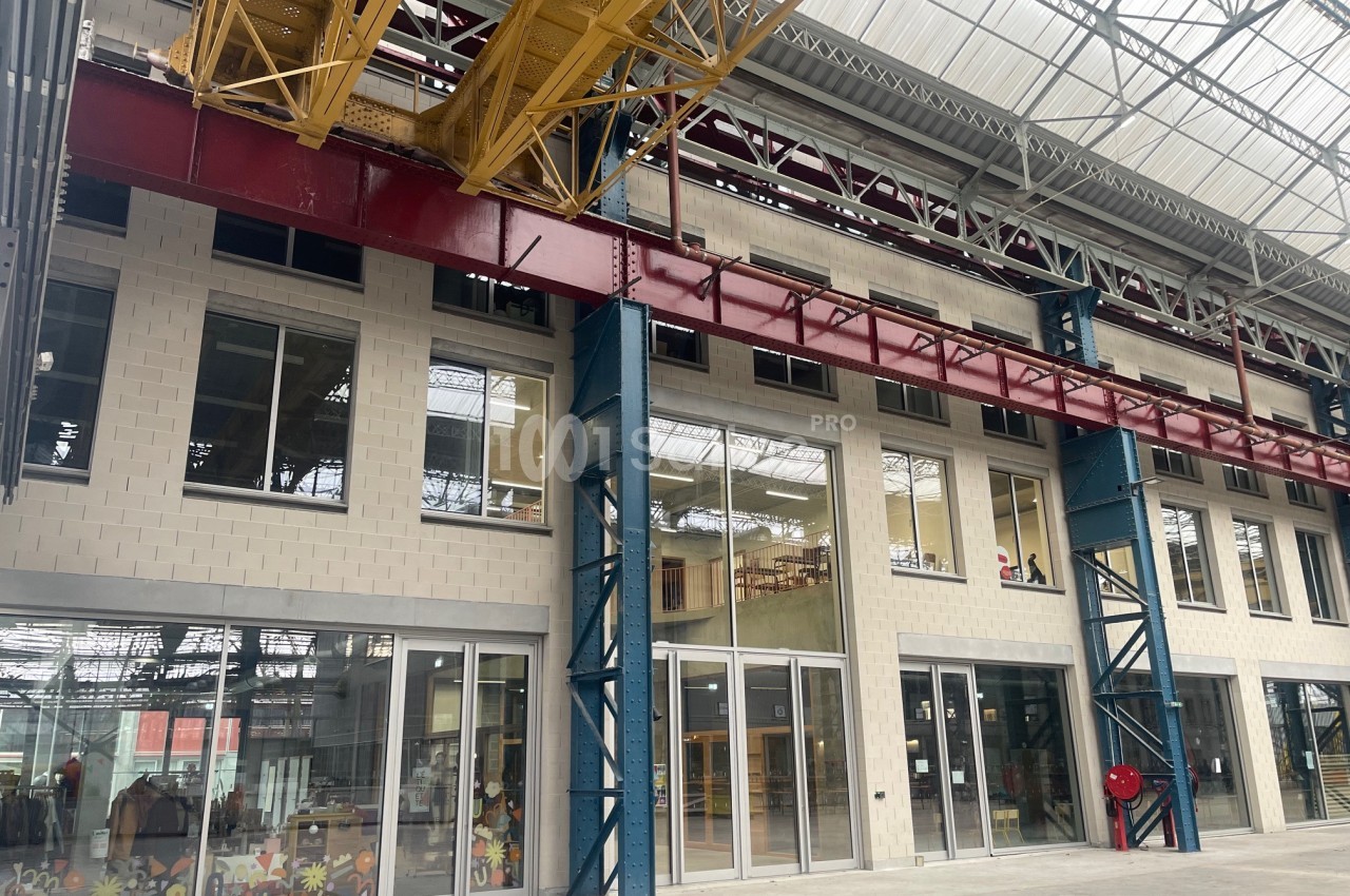 Structure industrielle rénovée avec de grandes baies vitrées, poutres métalliques colorées et plafond en verrière.