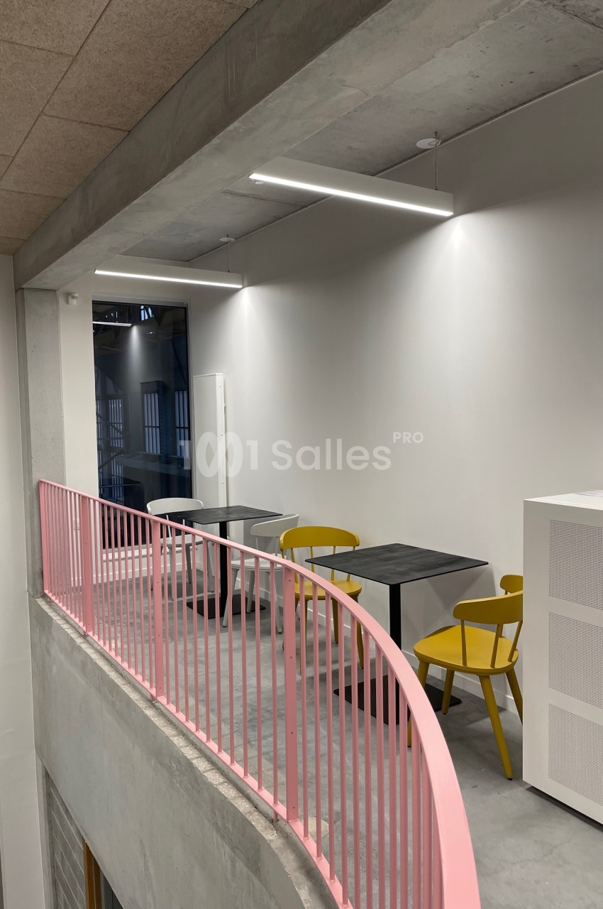 Espace mezzanine avec tables noires, chaises jaunes et rambarde rose, éclairé par des lampes suspendues.