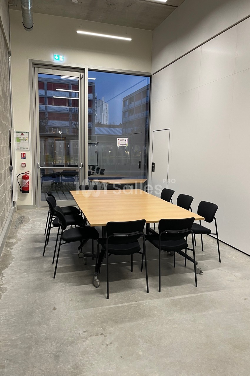 Salle de réunion avec une table rectangulaire en bois entourée de chaises noires, située près d'une grande baie vitrée.