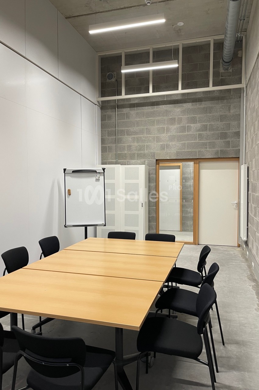 Salle de réunion avec table en bois, chaises noires, tableau blanc et murs en béton et panneaux blancs.