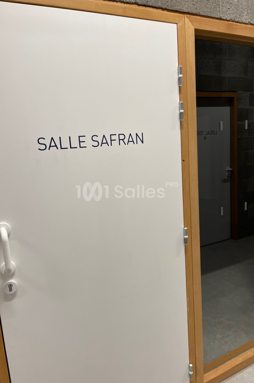 Porte blanche avec l'inscription ’Salle Safran’, encadrée de bois, donnant sur un espace intérieur en béton.