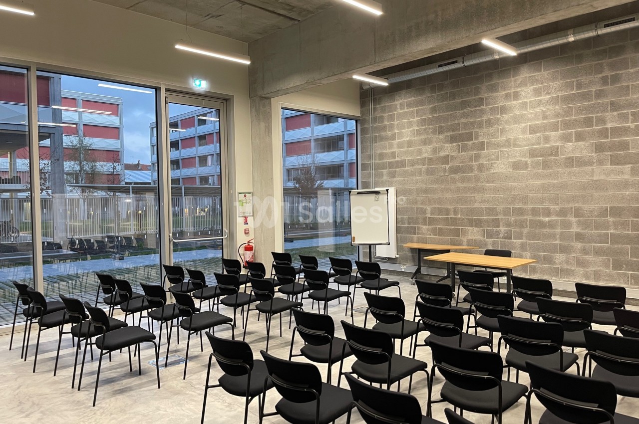 Salle de conférence vide avec des rangées de chaises noires, un tableau blanc et une table, éclairée par des fenêtres.