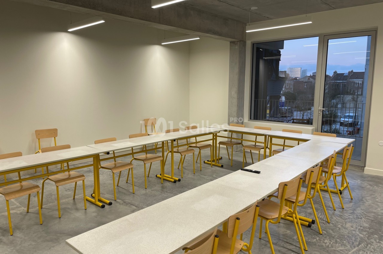 Salle de réunion lumineuse avec tables disposées en U, chaises en bois et grande fenêtre donnant sur un paysage urbain.