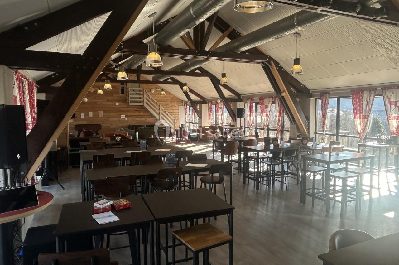 Salle de restaurant lumineuse avec poutres apparentes, grandes fenêtres, tables en bois et chaises hautes.