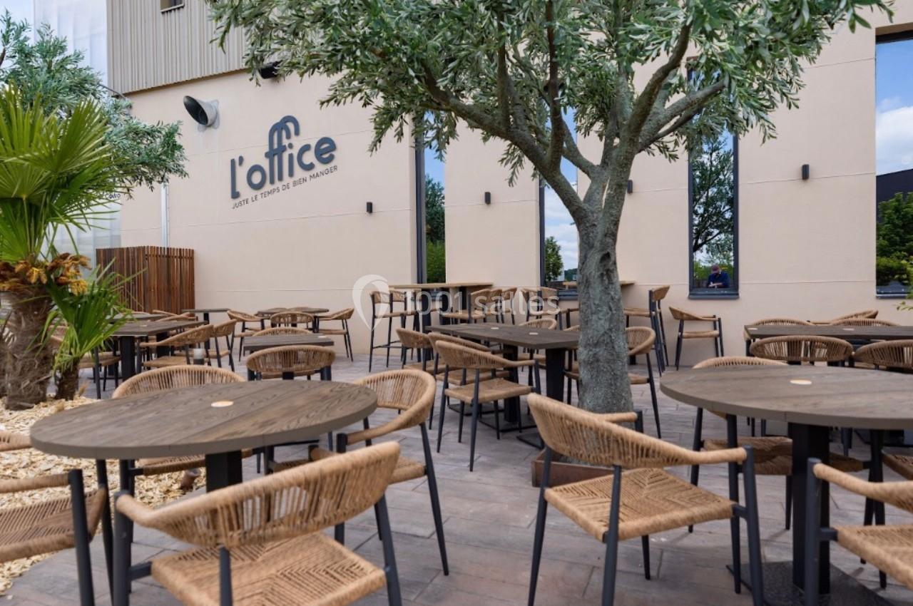 Terrasse extérieure avec tables et chaises en bois sous un olivier, devant un bâtiment beige marqué ’l'office’.