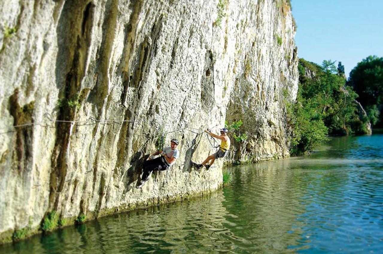 Deux personnes escaladent une paroi rocheuse au-dessus d'une rivière, équipées de harnais et de cordes.