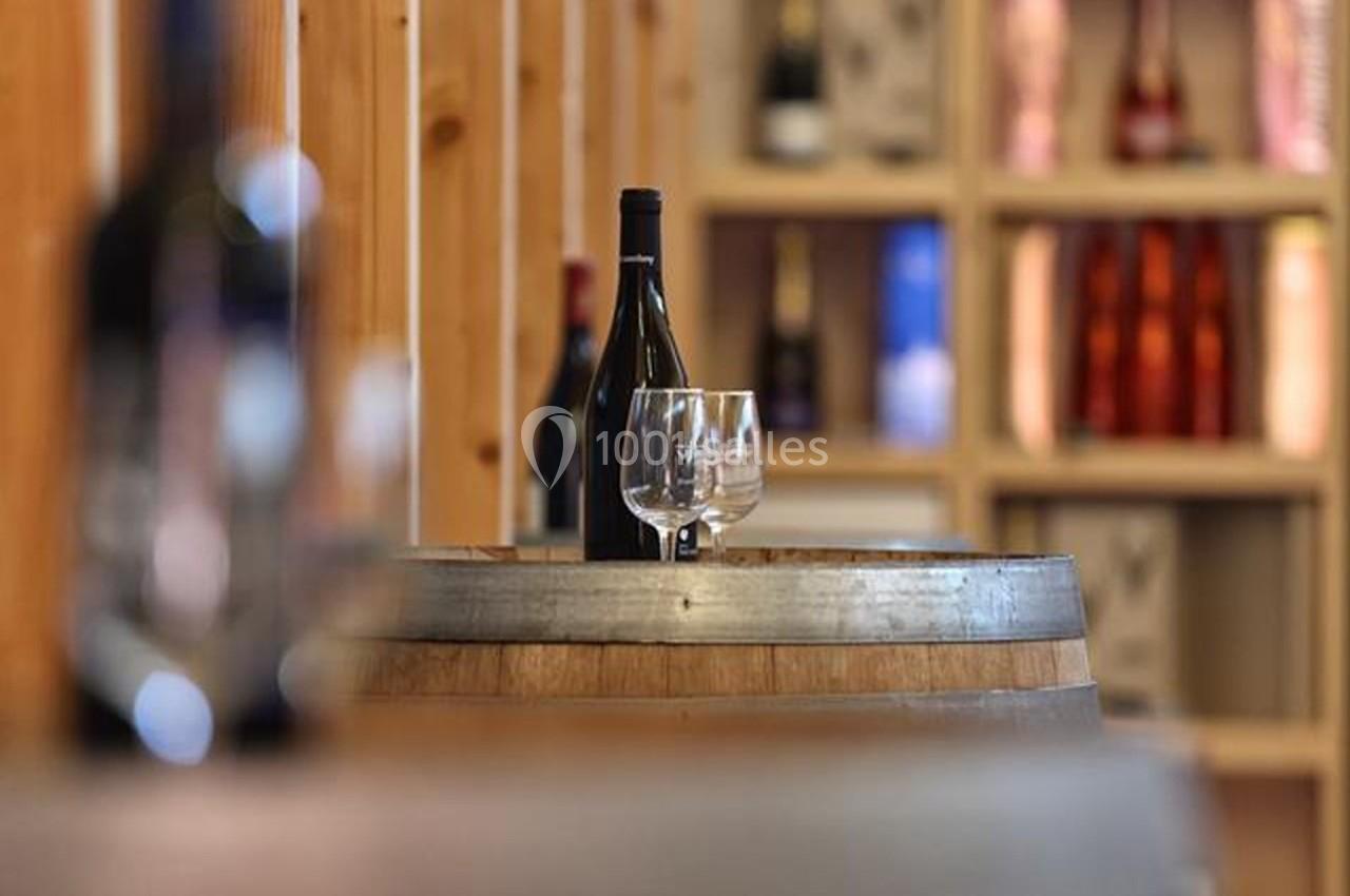 Bouteille de vin et deux verres posés sur un tonneau en bois, avec des étagères de bouteilles en arrière-plan flou.