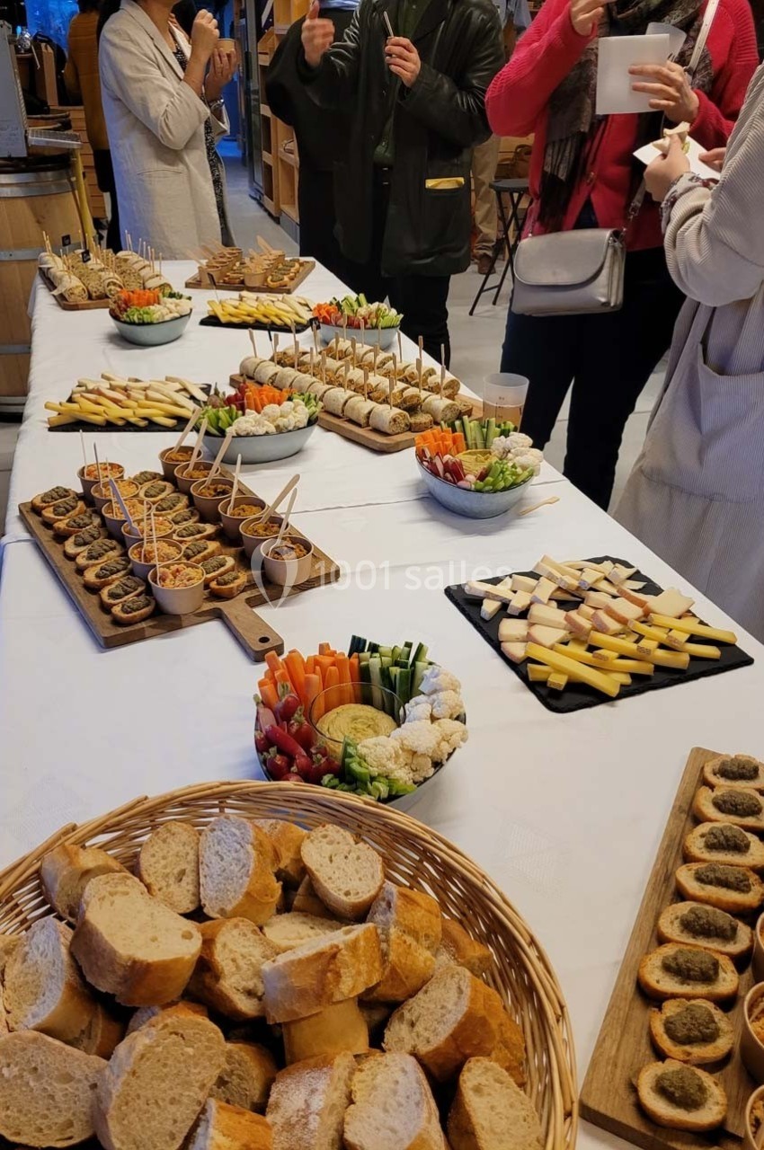 Table garnie de pains, fromages, légumes, tartes et amuse-bouches, entourée de personnes discutant debout.