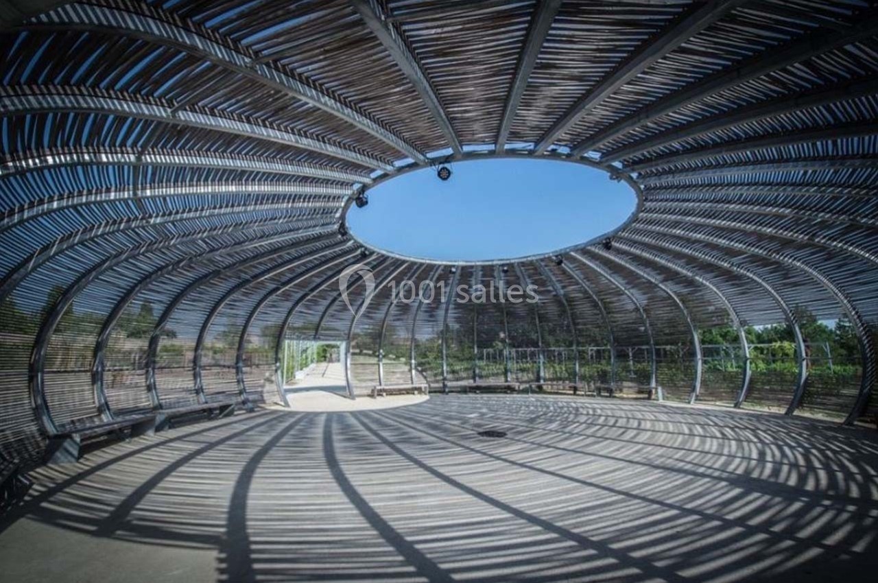 Structure circulaire en bois avec un toit ajouré, projetant des ombres sur le sol, entourée de végétation.