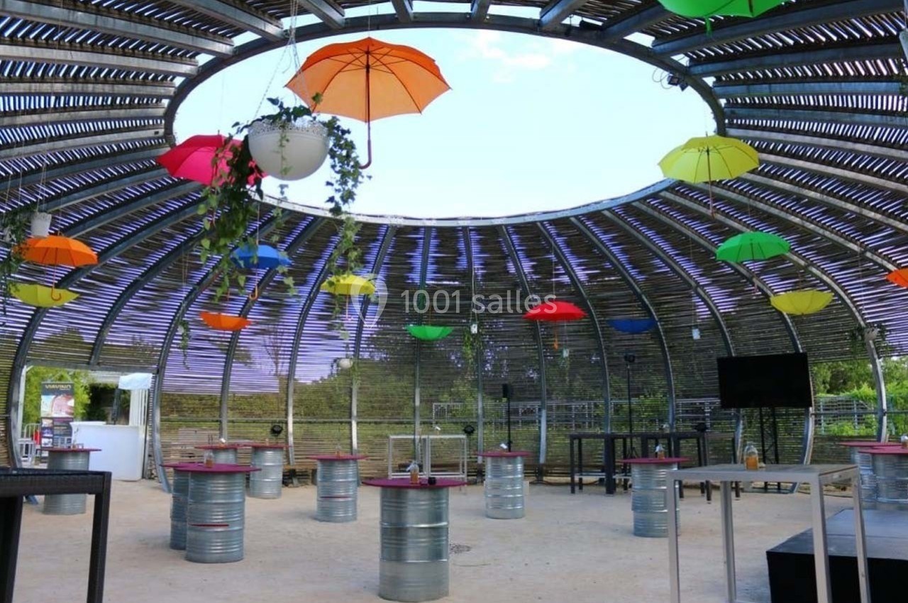 Structure circulaire en bois avec des tables en tonneaux et des parapluies colorés suspendus au plafond.