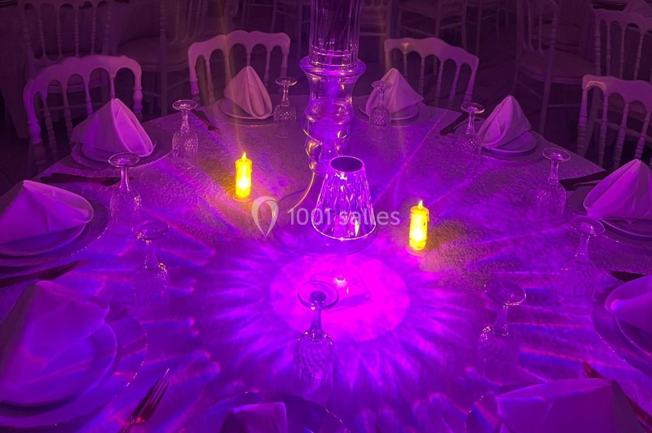 Table ronde élégamment dressée avec des serviettes pliées, des verres en cristal et un éclairage violet tamisé.
