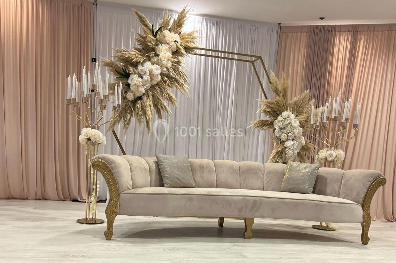 Canapé beige avec accoudoirs dorés, décoré de fleurs blanches, pampas et chandeliers sur fond de rideaux clairs.
