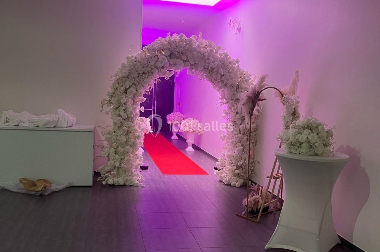 Arche florale blanche décorative avec tapis rouge menant à une porte, dans une salle éclairée par une lumière violette.