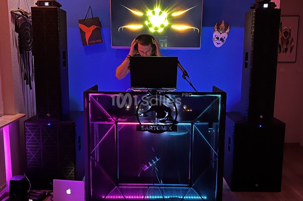 Un DJ mixe de la musique derrière une table lumineuse entourée de haut-parleurs dans une pièce décorée.