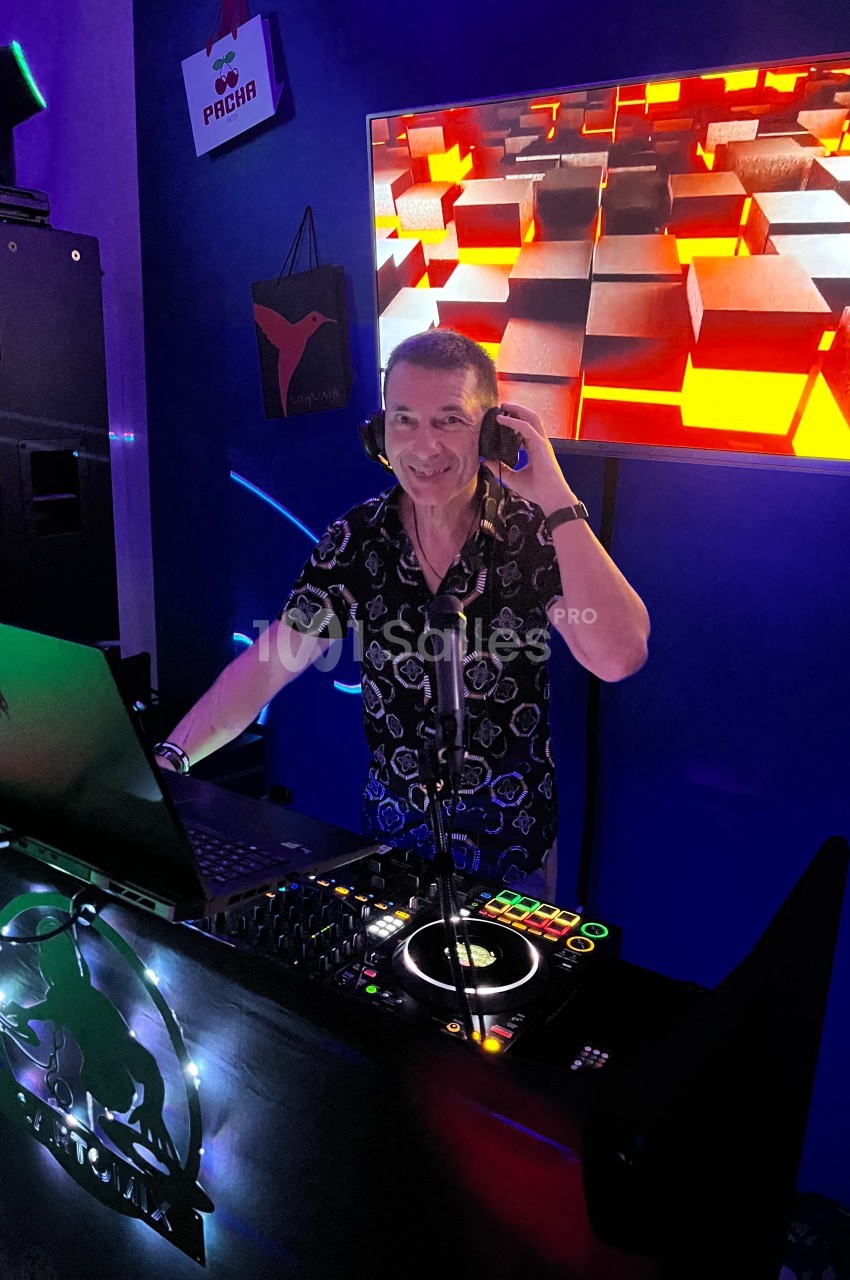 Un DJ souriant mixe de la musique dans un espace sombre, éclairé par des lumières colorées et un écran lumineux.
