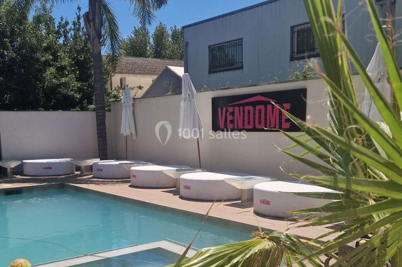 Piscine extérieure avec transats ronds, parasols blancs et panneau ’Vendôme’ sur un mur en arrière-plan.