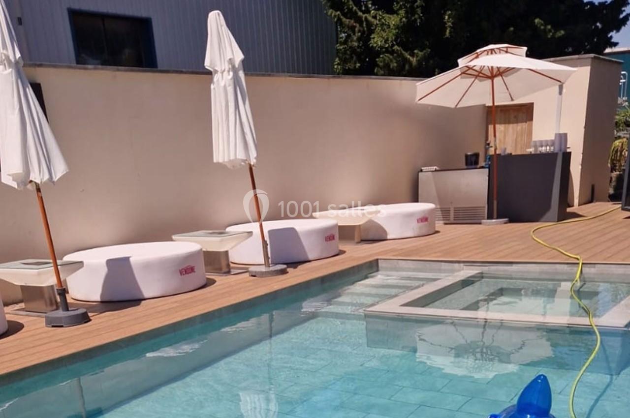 Terrasse avec piscine, transats blancs, parasols fermés et un bar en arrière-plan sous un ciel dégagé.