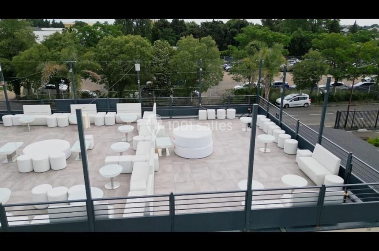 Terrasse aménagée avec mobilier blanc moderne, entourée de verdure et donnant sur un parking en arrière-plan.
