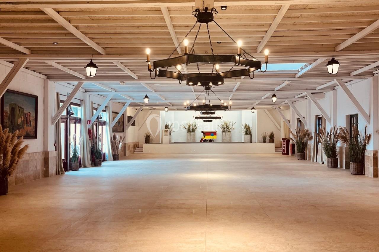 Grande salle vide avec un sol en pierre, plafond en bois avec poutres apparentes et lustres suspendus, décorée de plantes.