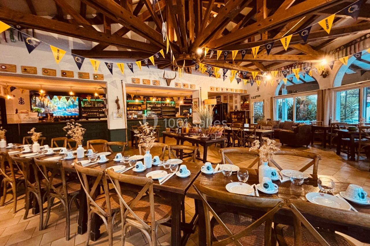 Salle de restaurant décorée avec des guirlandes, tables en bois dressées avec vaisselle et vue sur un bar éclairé.