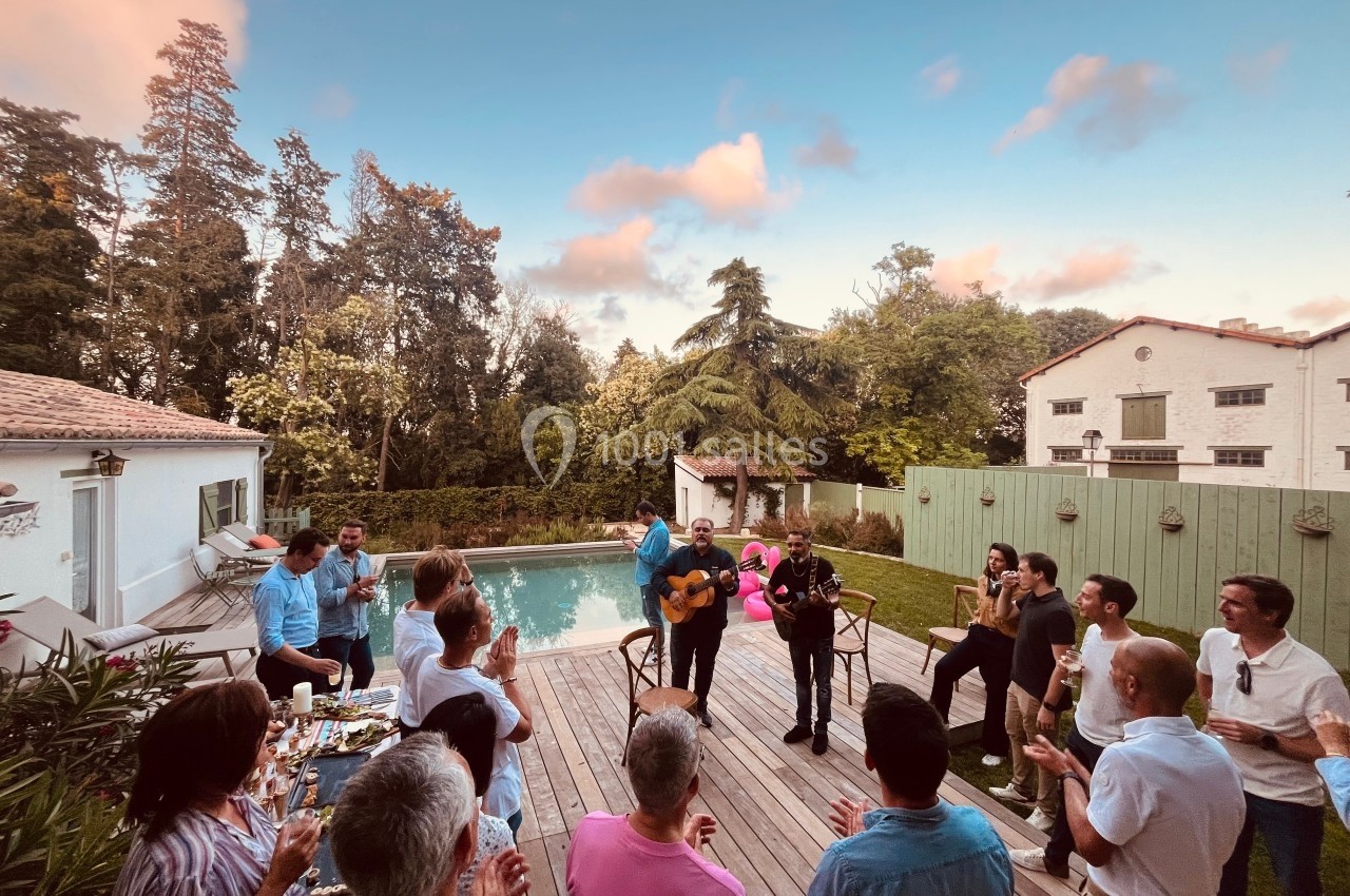 Groupe de personnes réunies autour de musiciens jouant près d'une piscine dans un jardin au coucher du soleil.