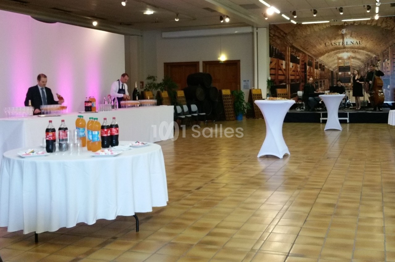 Salle de réception avec tables dressées, boissons, personnel de service et musiciens sur une scène en arrière-plan.