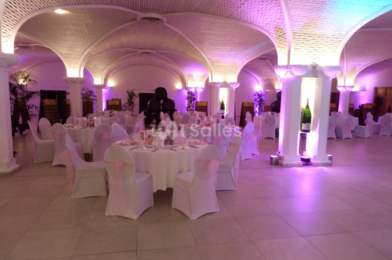 Salle de réception élégante avec tables rondes dressées, chaises blanches et éclairage violet sous des voûtes en briques.