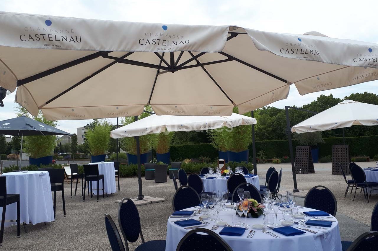 Tables dressées avec nappes blanches et chaises bleues sous de grands parasols dans un espace extérieur arboré.