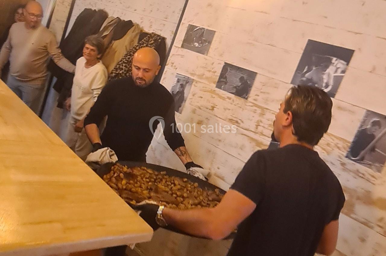 Deux hommes portent un grand plat rempli de châtaignes dans un intérieur avec des murs clairs et des photos accrochées.