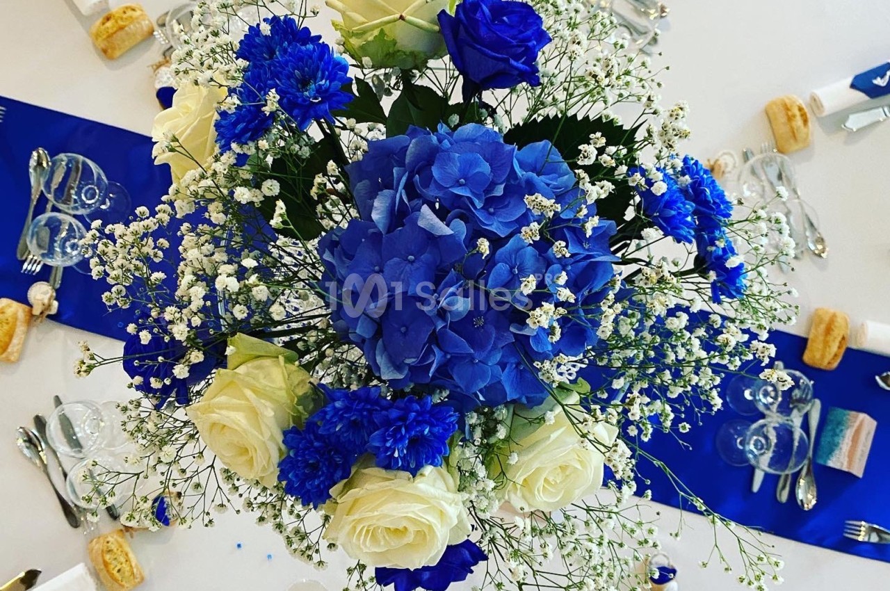 Bouquet de fleurs blanches et bleues au centre d'une table dressée avec des couverts et des décorations assorties.