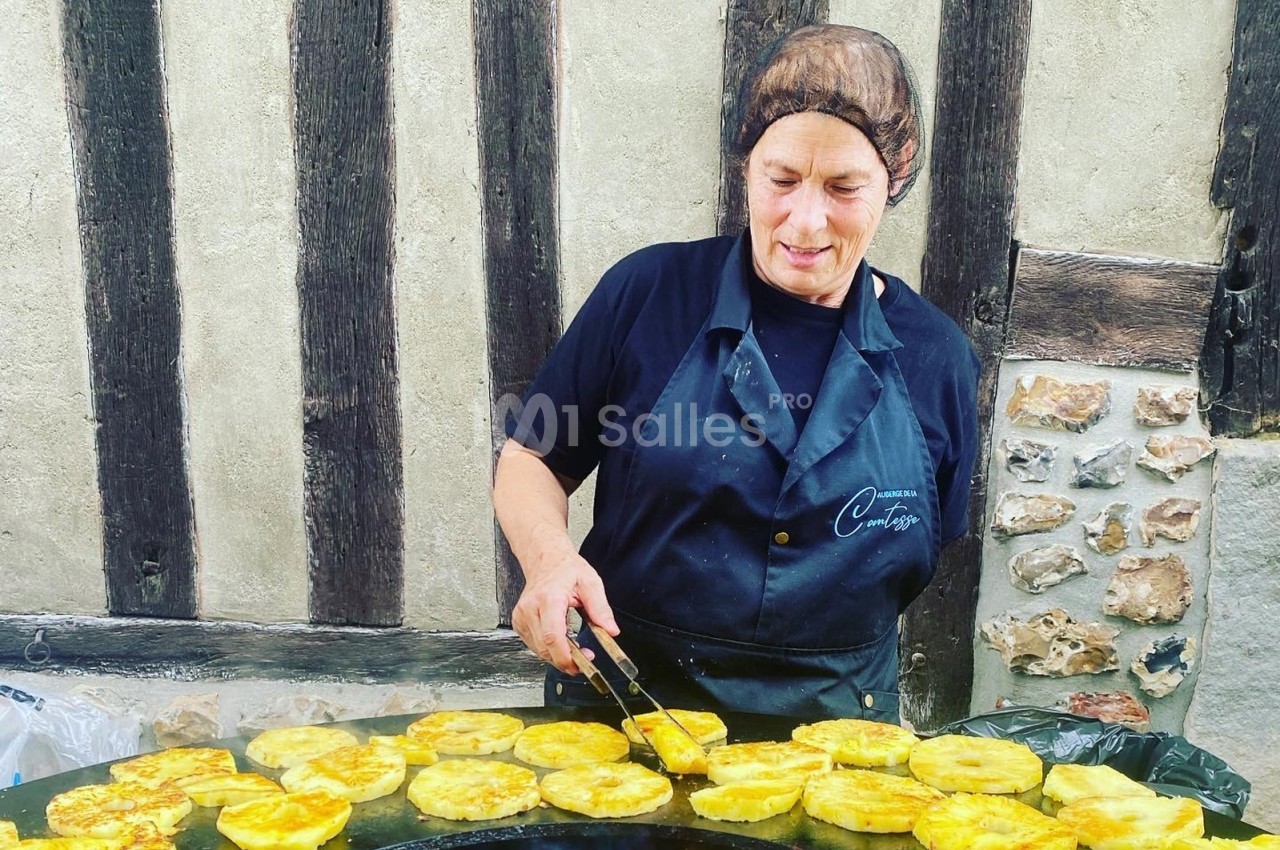 Femme en tablier noir cuisant des tranches de légumes dorés sur une grande plaque chauffante devant un mur à colombages.