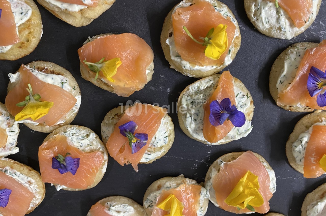 Canapés au saumon fumé garnis de fleurs comestibles et de fromage frais aux herbes, disposés sur une ardoise.