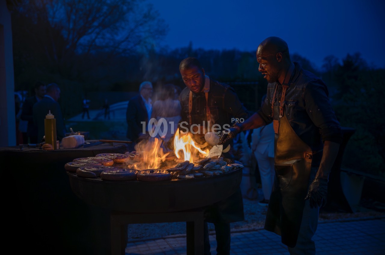 Deux hommes cuisinent des aliments sur un grand barbecue en plein air, éclairé par les flammes, à la tombée de la nuit.
