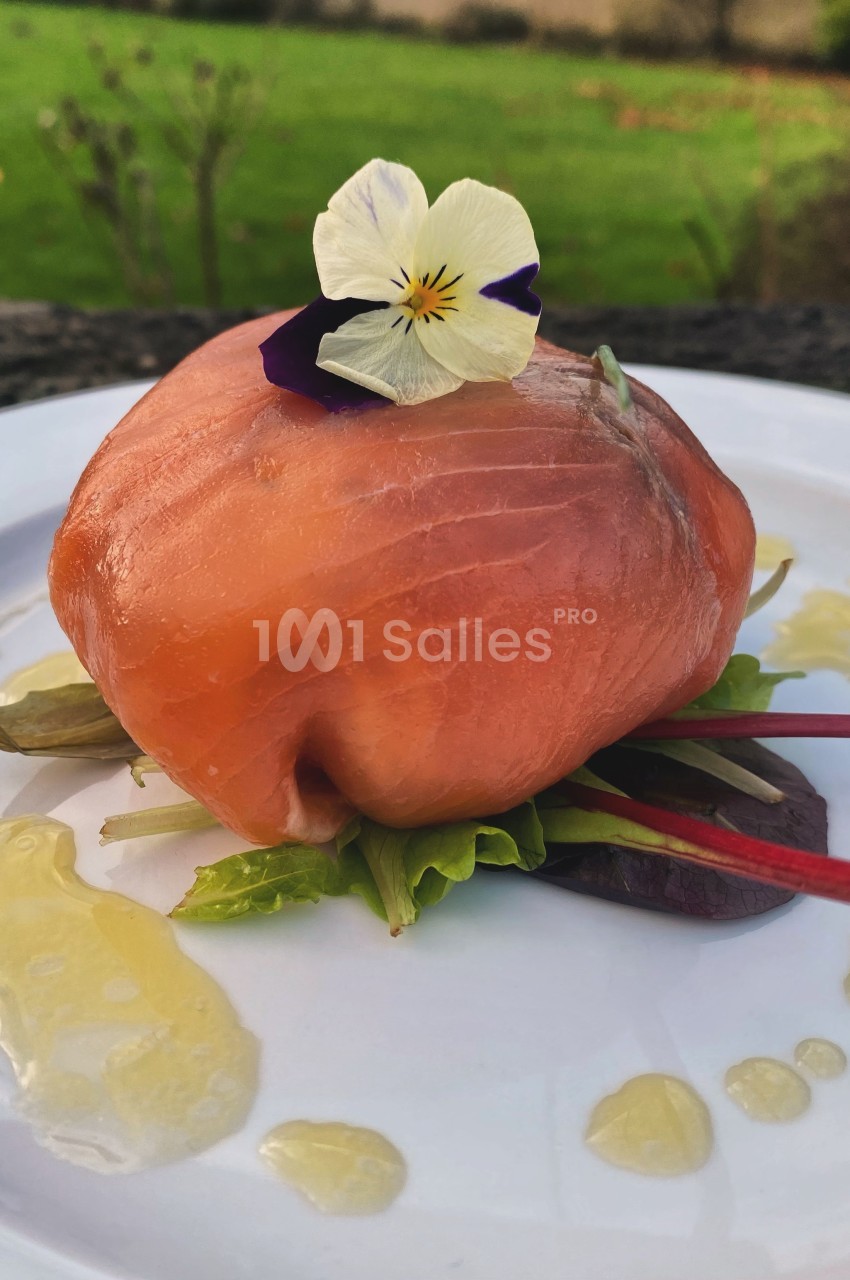Ballotine de saumon fumé garnie de jeunes pousses, décorée d'une fleur comestible, servie avec un filet d'huile.