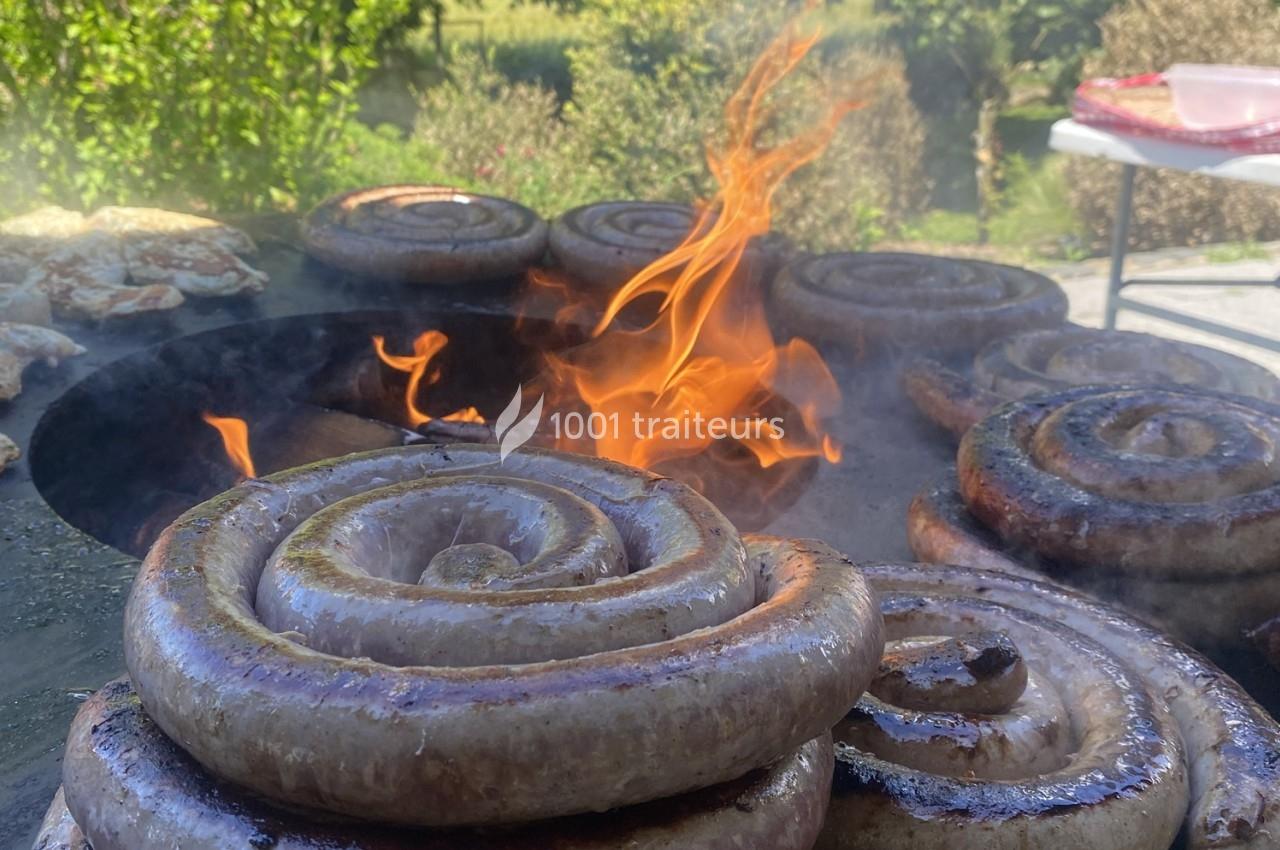 Saucisses en spirale grillant sur un barbecue avec des flammes vives en arrière-plan, dans un cadre extérieur verdoyant.