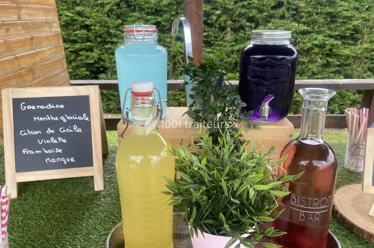 Boissons variées en carafes et bouteilles sur une table en bois, avec une ardoise listant les saveurs disponibles.