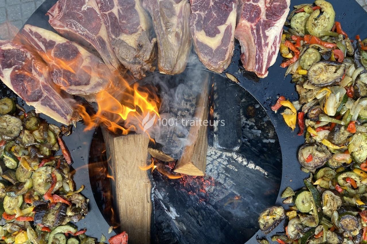 Viandes et légumes grillés disposés autour d'un feu de bois sur une plaque de cuisson circulaire.
