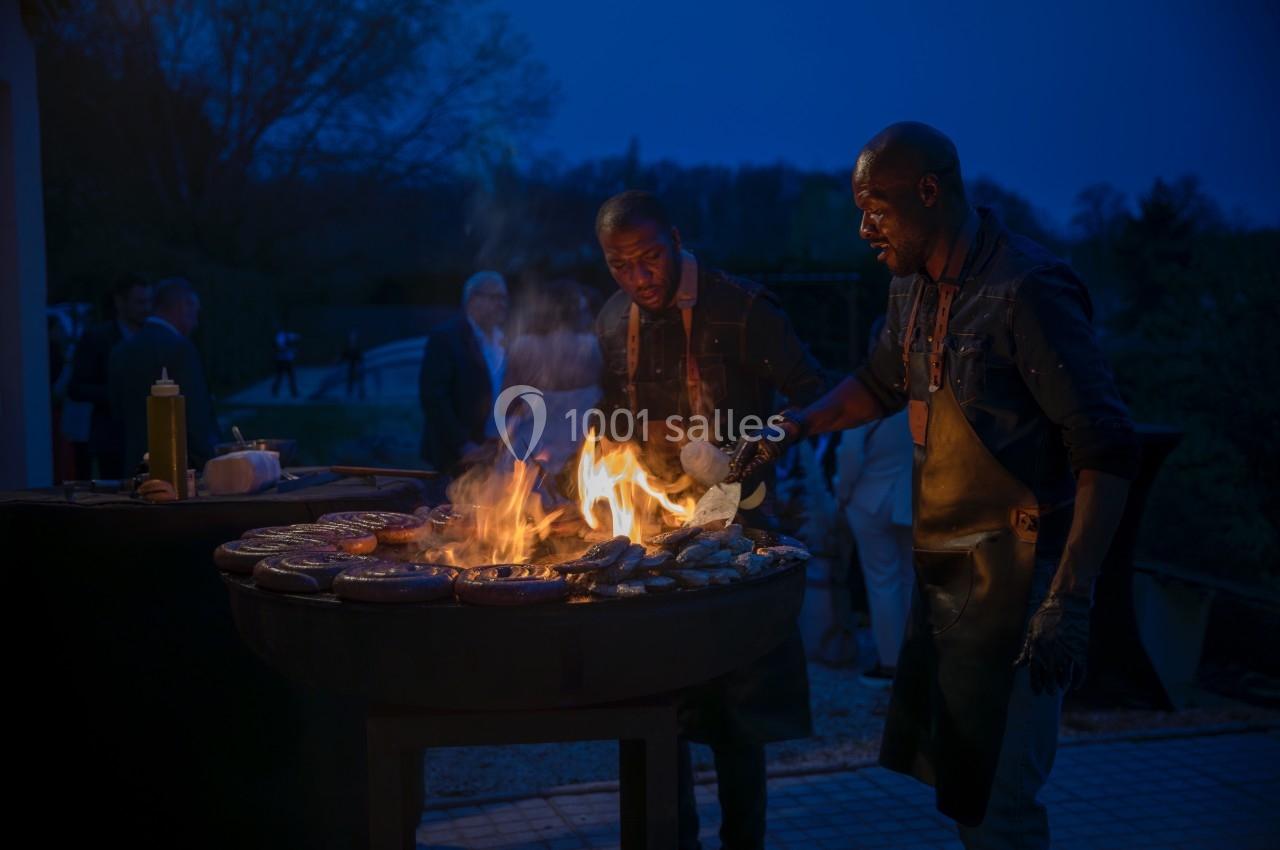 Deux hommes cuisinent des aliments sur un grand barbecue en plein air, éclairé par les flammes, à la tombée de la nuit.