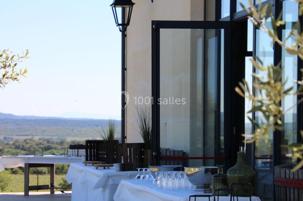Tables dressées en extérieur avec des verres et des bouteilles, près d'un bâtiment avec vue sur un paysage naturel.