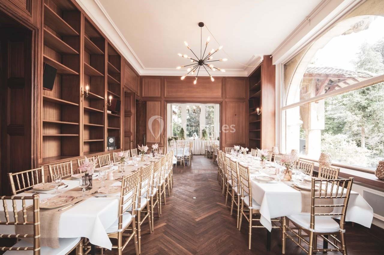 Salle élégante avec tables dressées, chaises dorées, parquet en bois, étagères murales et grande baie vitrée donnant sur un…