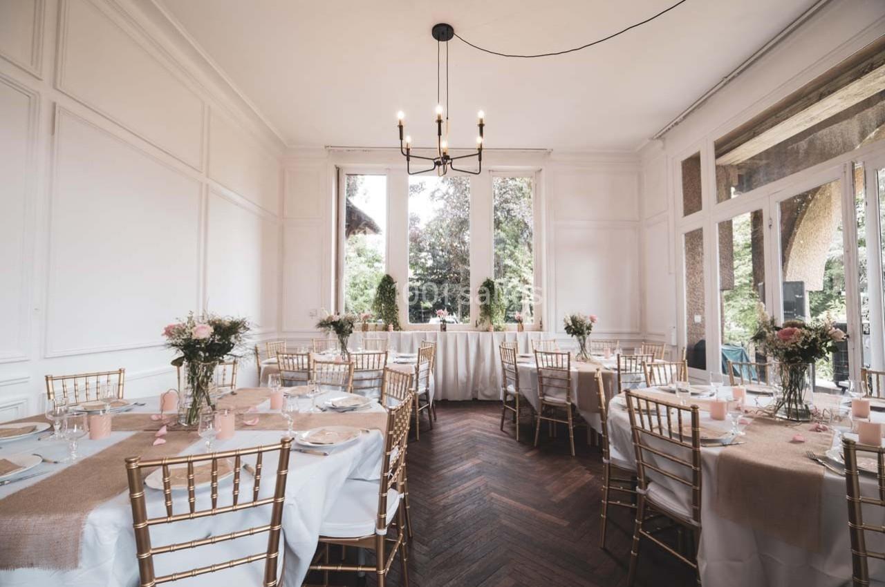 Salle lumineuse décorée pour un événement, avec tables rondes, chaises dorées et grandes fenêtres donnant sur un jardin.
