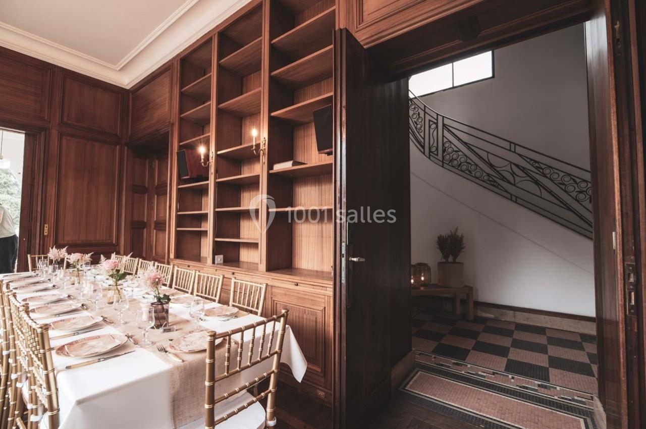 Salle à manger élégante avec table dressée et chaises dorées, ouverte sur un hall avec escalier en fer forgé.